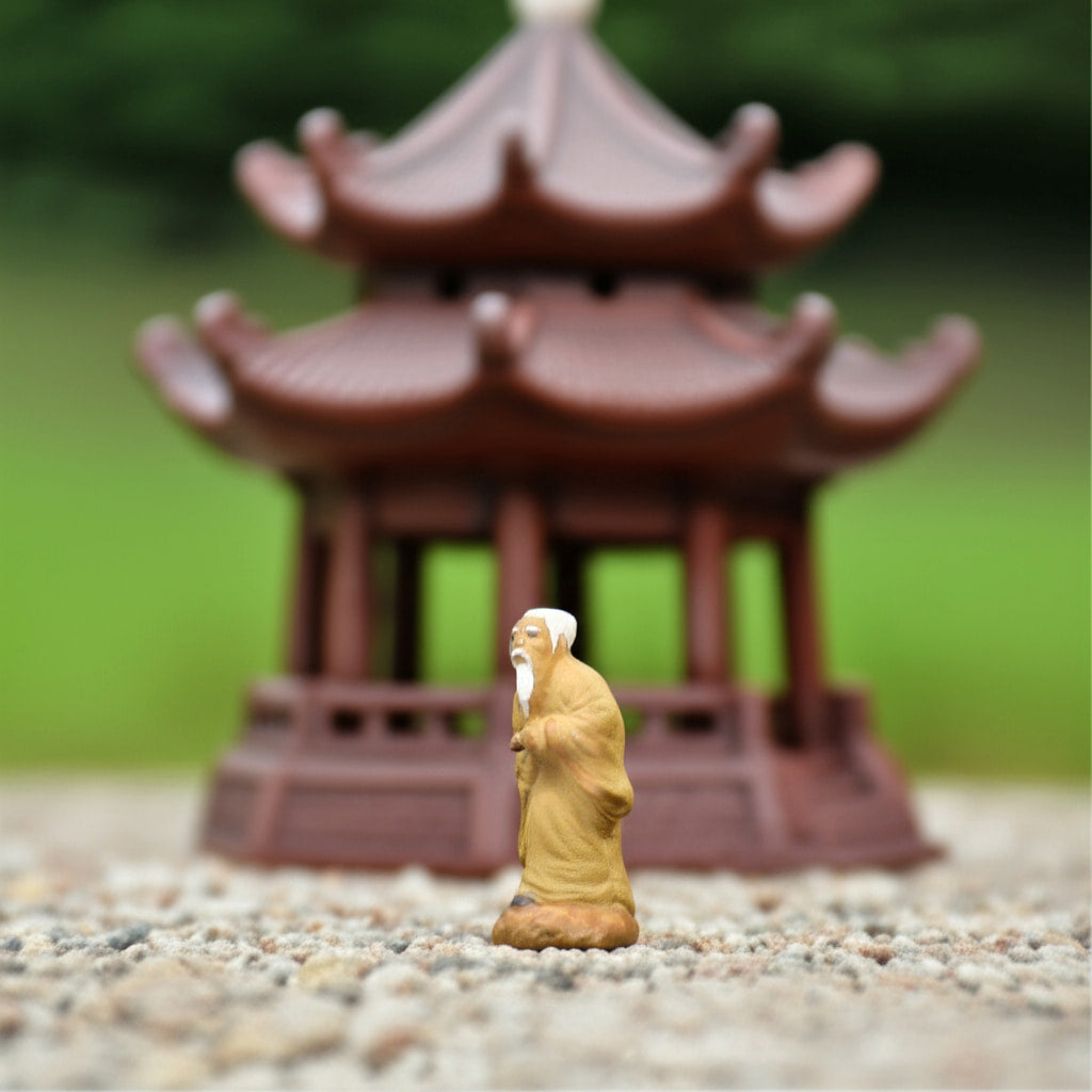 A wise old man, one of our bonsai figurines, stands on gravel before a blurry miniature pagoda.