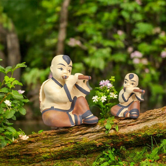 Two ceramic figurines of boys playing flutes sit on a mossy log. One figurine is larger than the other.