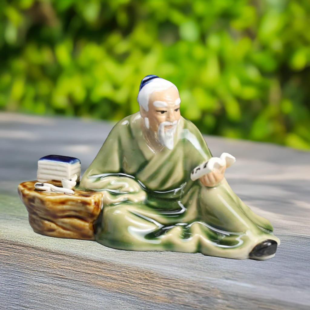A ceramic mudman with books figurine sits on a wooden surface, ideal for bonsai decor, with a green background.