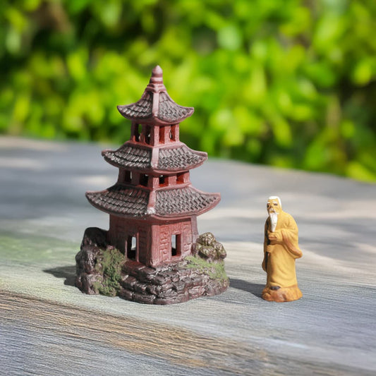 A miniature red pagoda and a mudman figurine are placed on a wooden surface with a green, leafy background. Suitable for Japanese terrarium decor.