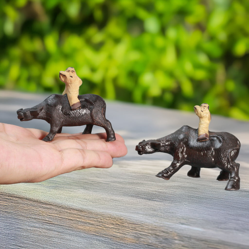 Two dark brown ceramic statues of a boy riding a water buffalo are held in a hand.