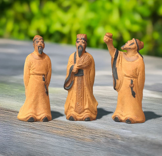 Three small unglazed ceramic figures standing on a wooden surface. The figures depict Zhuge Liang holding a fan, Li Bai with his hands clasped, and Li Bai raising a cup.