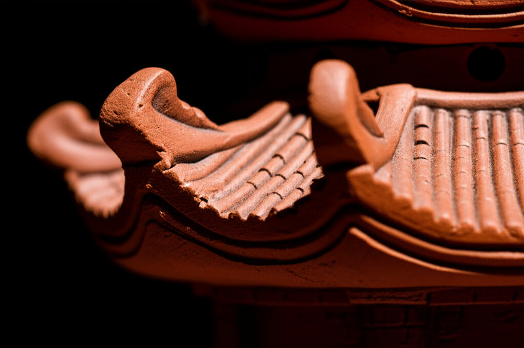 A close-up of a reddish-brown ceramic pagoda statue's detailed, tiled roof against a black background.