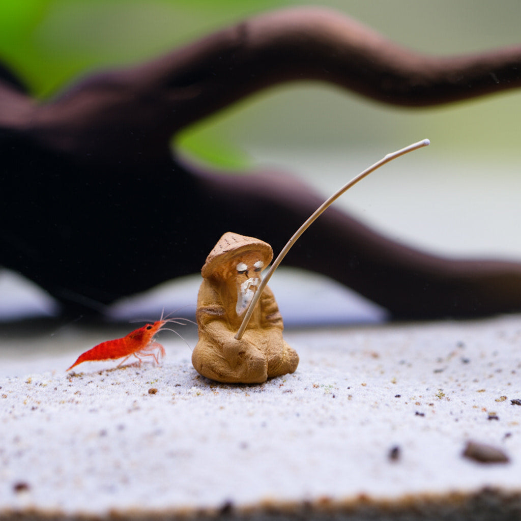 This small fisherman figure sits on sand near a shrimp, a unique piece of underwater fisherman decor.