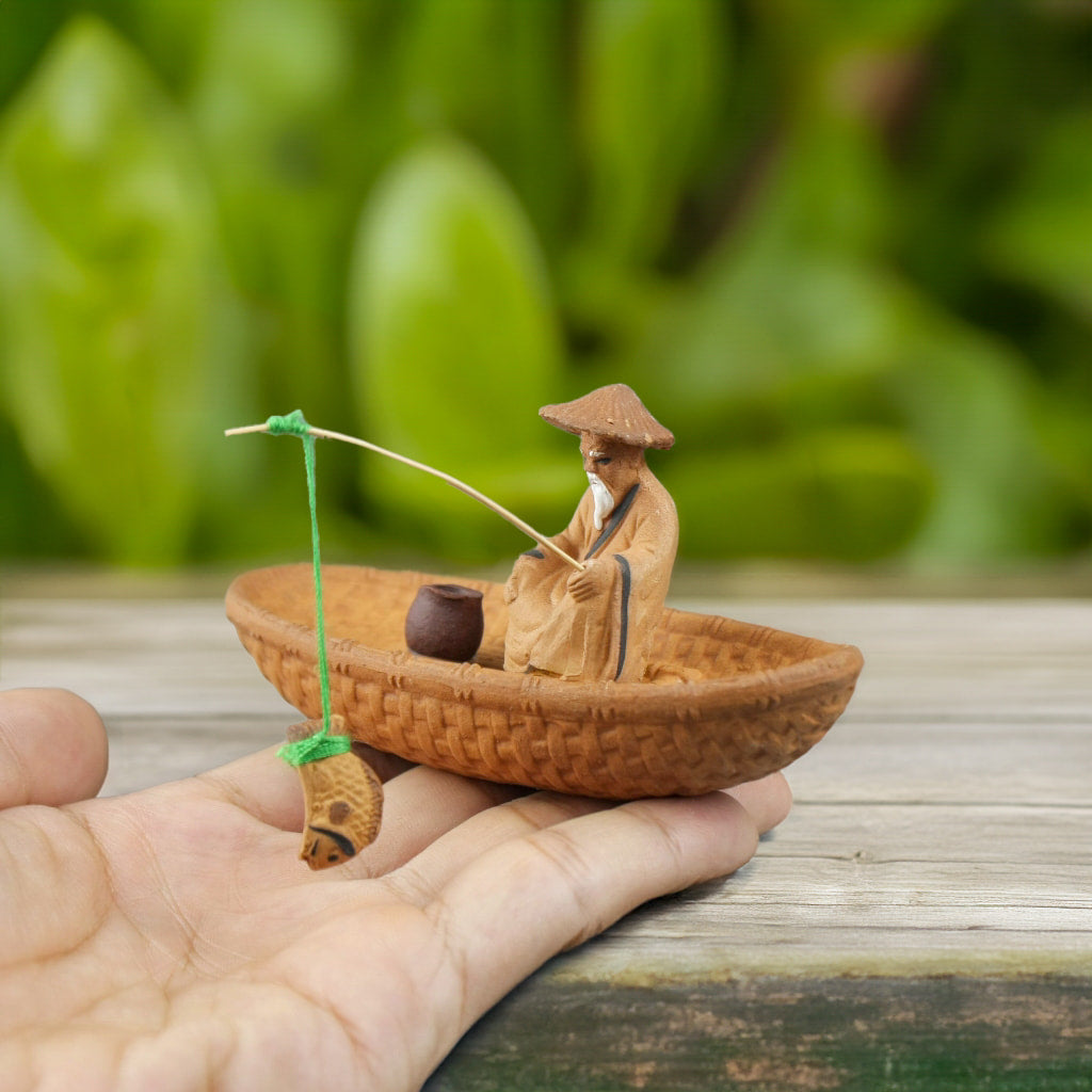 Hand holding a fisherman figurine in a boat, showing its small size for bonsai decorations.