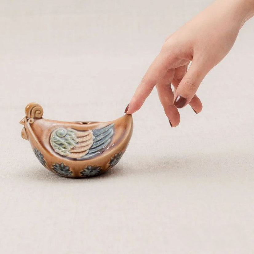 A hand touches a brown Vietnamese pottery rooster, an artisan chicken statue for shelf from Vietnam