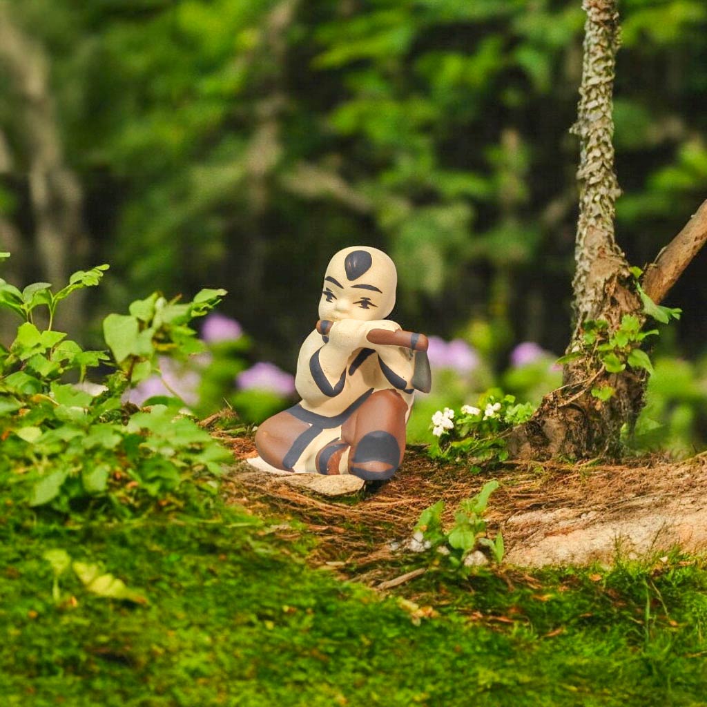 A small ceramic figurine of a boy playing a flute sits on a mossy area, surrounded by green plants.