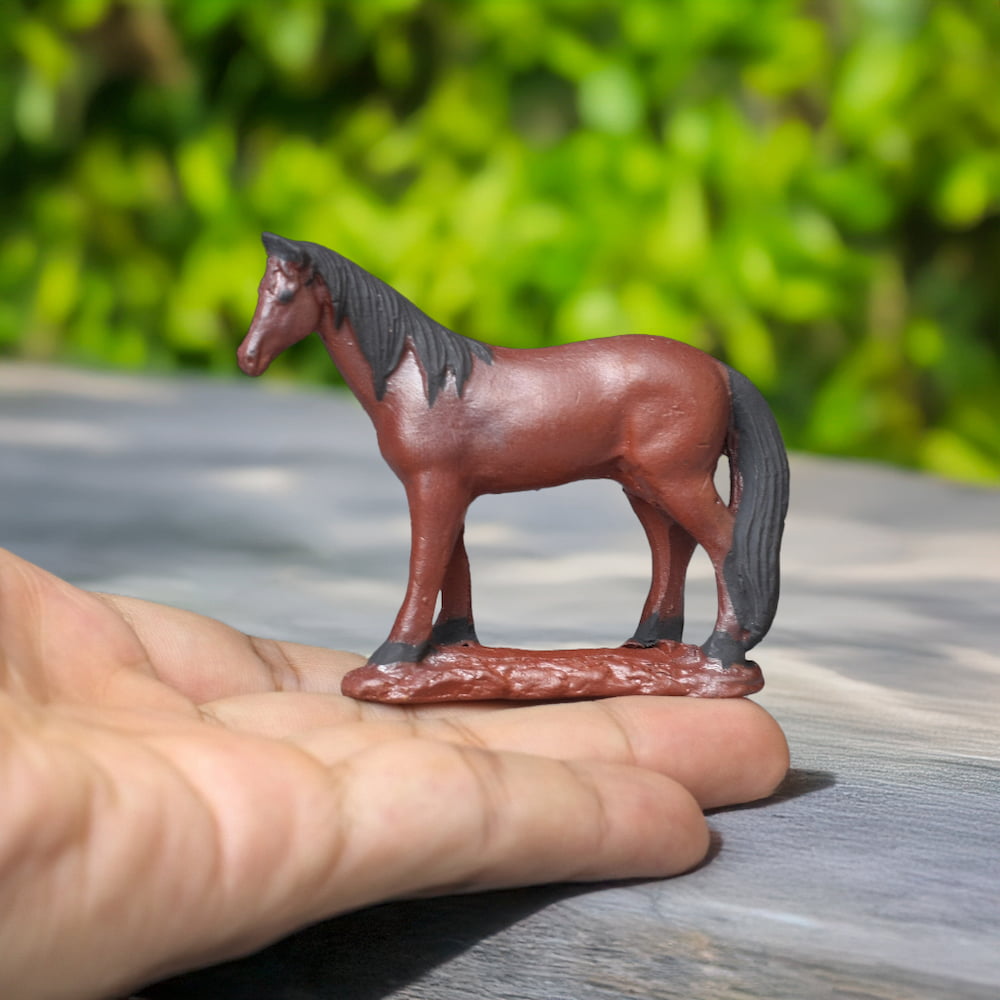 A small, brown ceramic horse with a black mane and tail stands on a hand against a blurred green background.
