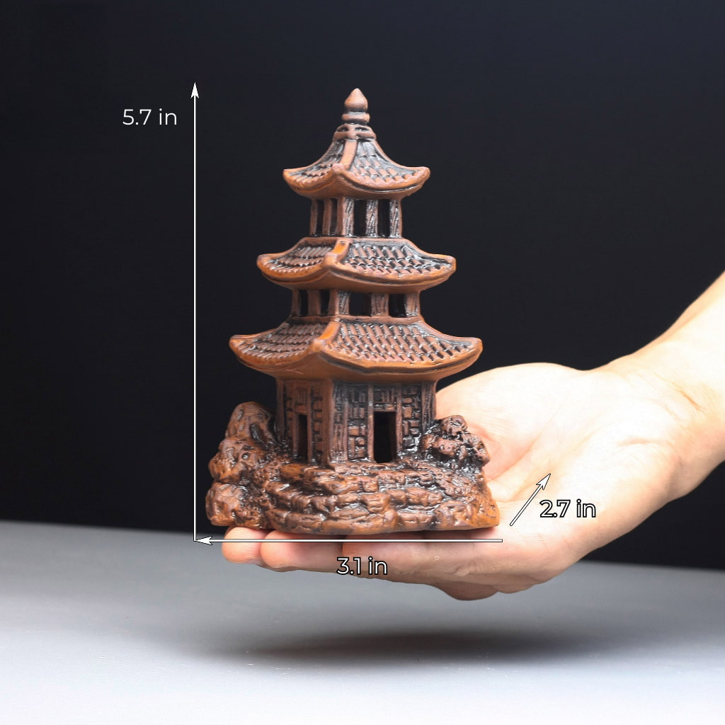 A small, brown ceramic pagoda sits on a hand. It has three tiers and a pointed roof. The base is textured to resemble a rocky outcrop.