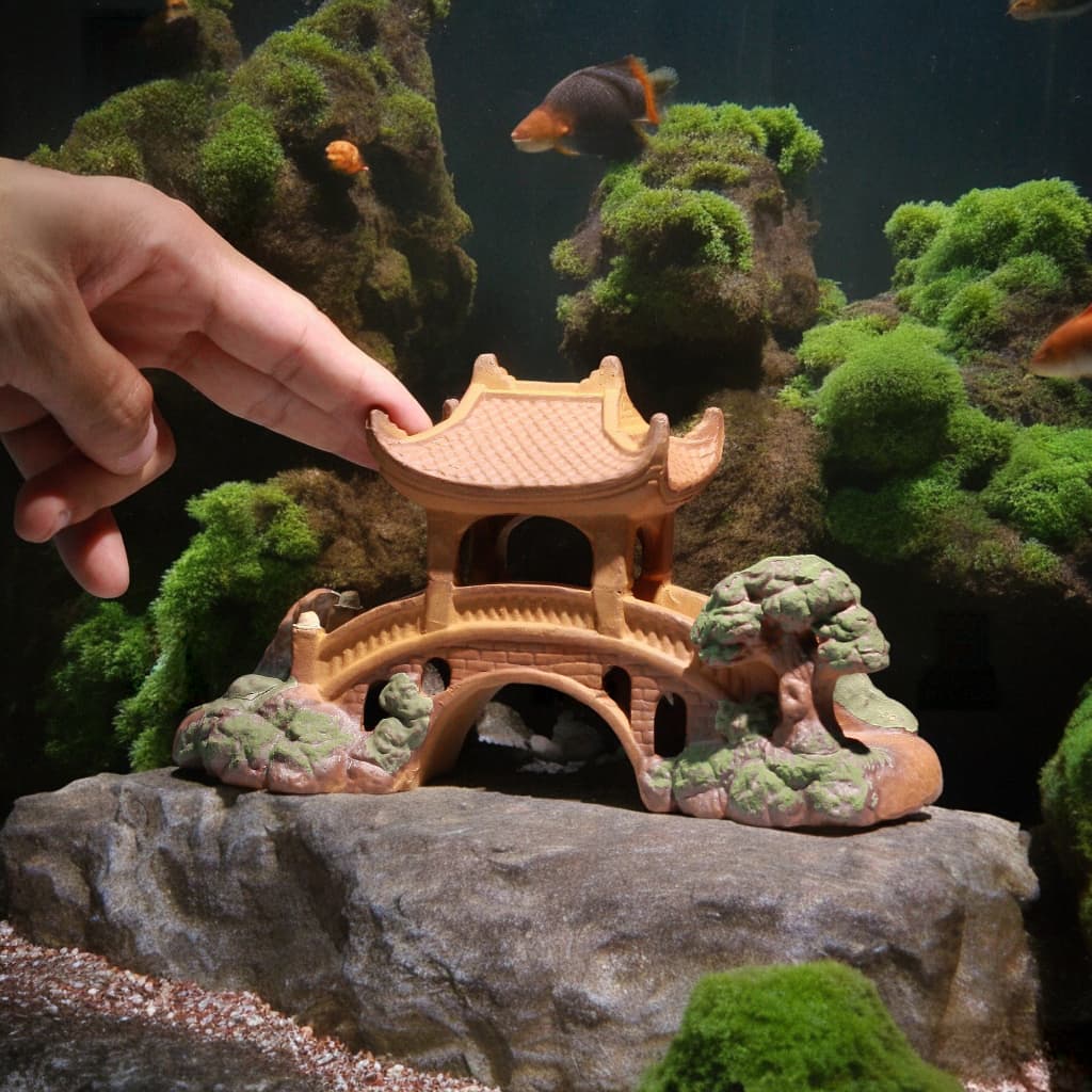 Hand places ceramic pavilion miniature bridge on rock. Aquarium decor, a fairy bridge style.