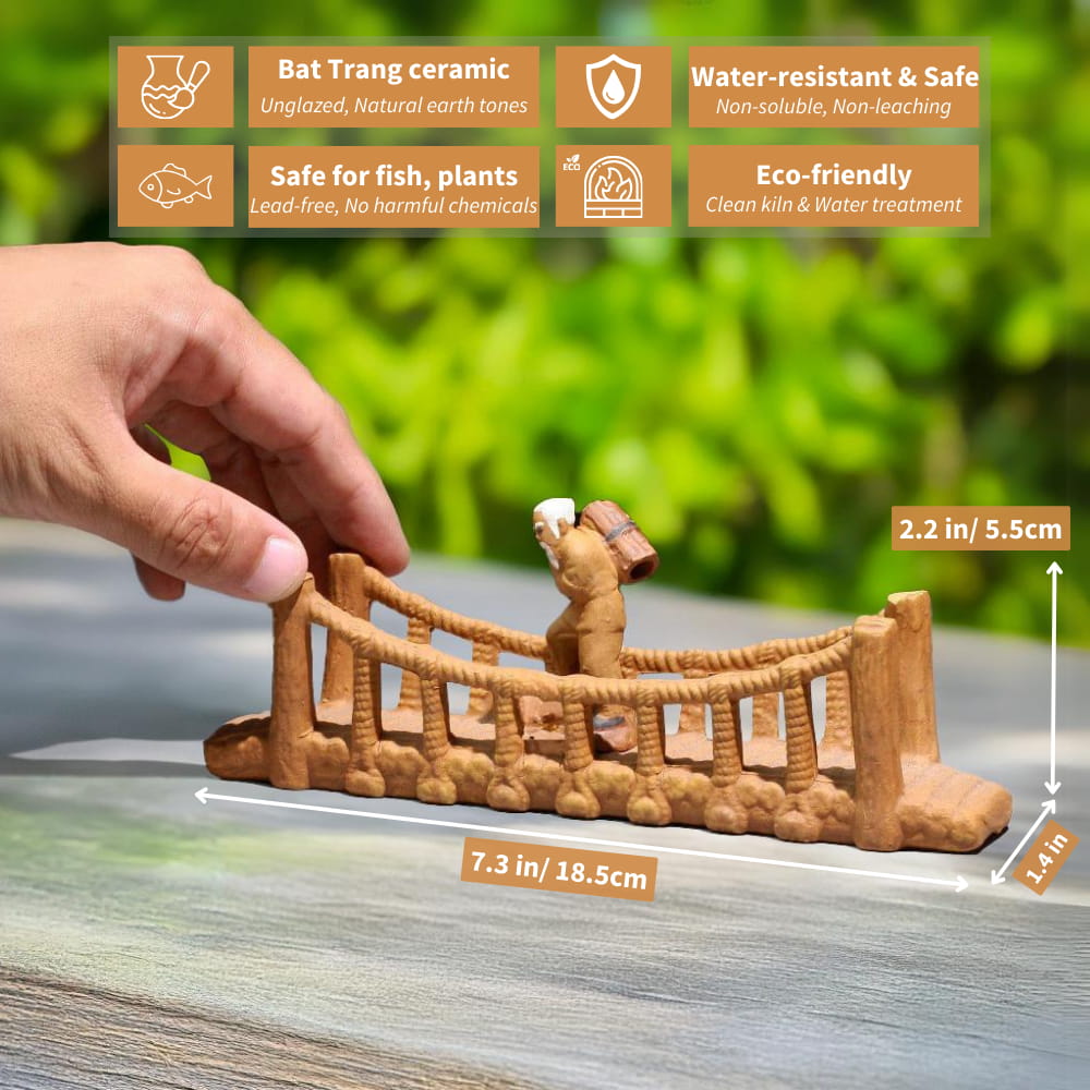 A small ceramic bridge with a rope railing, sitting on a wooden surface. A small figure of an old man is walking across the bridge. The bridge is unglazed and has a natural brown color.