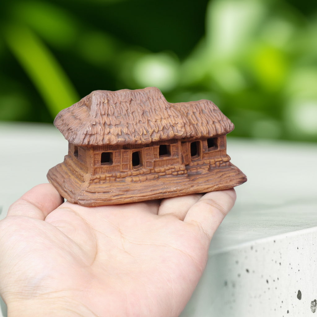 A small, brown thatched house figurine with a textured roof rests in an open hand. This miniature house figurines fits perfectly.