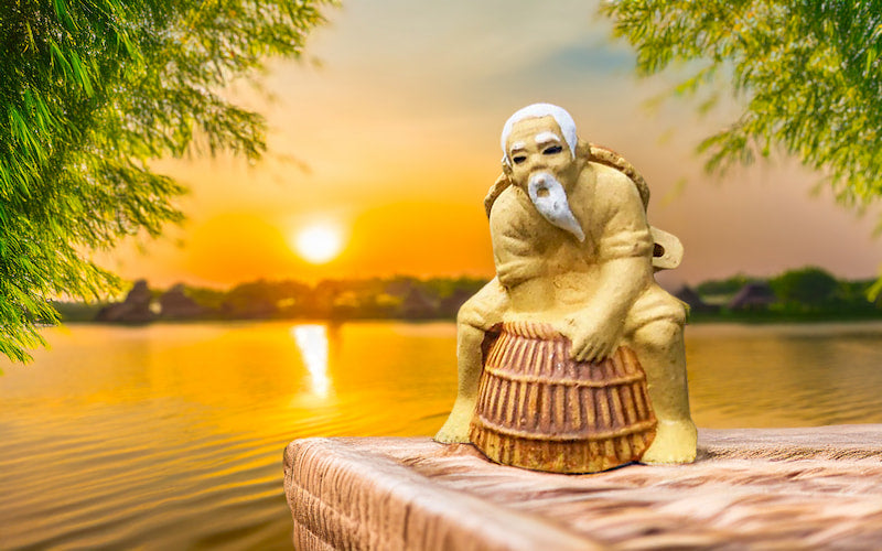 As the sun sets over a tranquil Vietnamese lake, casting a golden glow, a weathered mudman figurine sits serenely on a wooden perch, his basket trap at his side, evoking a sense of timeless tradition.