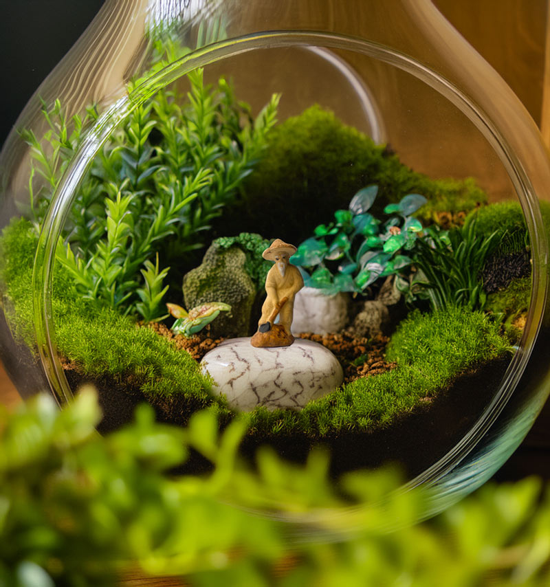 Inside a lush green terrarium, a miniature gardener figurine diligently works amidst delicate ferns and moss, adding a touch of life and tranquility to this miniature ecosystem.