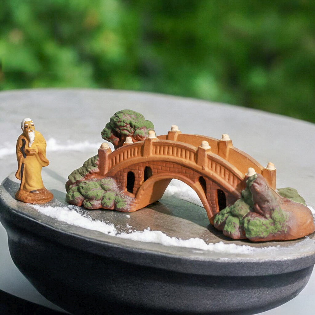 This small garden bridge, a fairy bridge, sits on a pot with a mudman figure beside it.