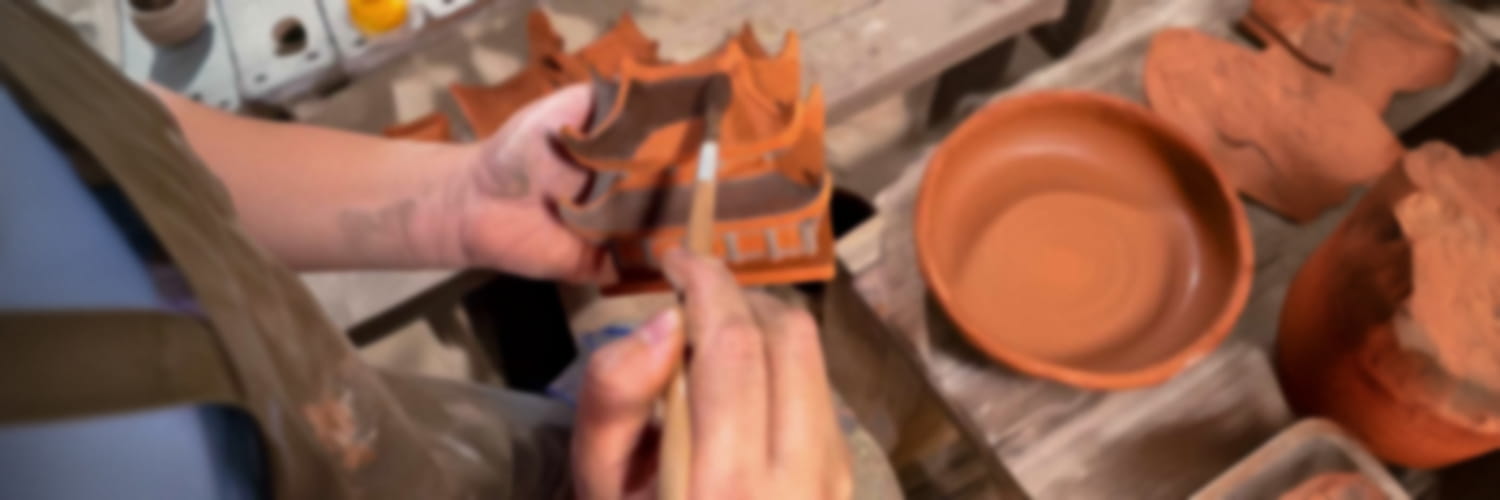 An artisan’s hands, stained with clay, meticulously paint intricate details onto a Vietnamese ceramic figurine. Surrounded by rustic terracotta bowls and the tools of their craft, they breathe life into each unique piece, carrying on the centuries-old tradition of Bat Trang ceramic art.