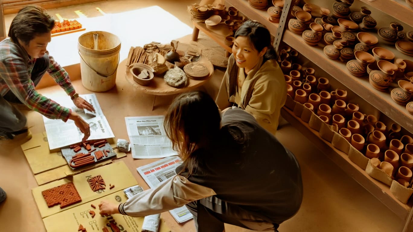 Inside the sunlit Duc Huy Pottery Workshop, a group of artisans and students gather around a table, their hands busy shaping intricate ceramic tiles. The warm atmosphere reflects The Viet Potter's commitment to fostering community and nurturing the next generation of Vietnamese ceramic artists.