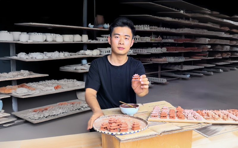 A skilled artisan at Duc Huy Pottery Workshop proudly displays a miniature ceramic pagoda, surrounded by shelves overflowing with unique, handcrafted Vietnamese ceramic figurines, ready to be shipped worldwide by The Viet Potter.