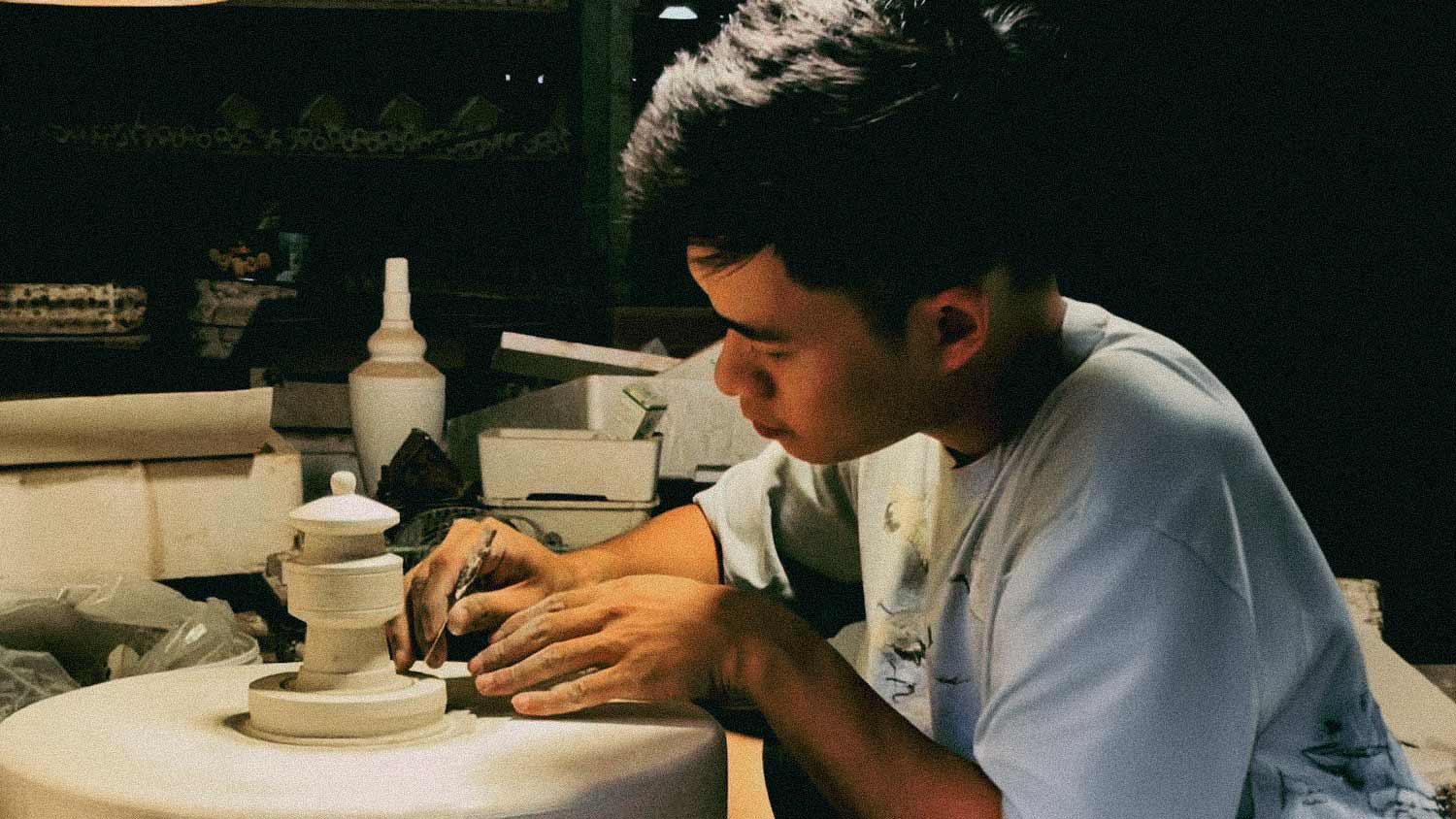 A skilled artisan at Duc Huy Pottery Workshop proudly displays a miniature ceramic pagoda, surrounded by shelves overflowing with unique, handcrafted Vietnamese ceramic figurines, ready to be shipped worldwide by The Viet Potter.