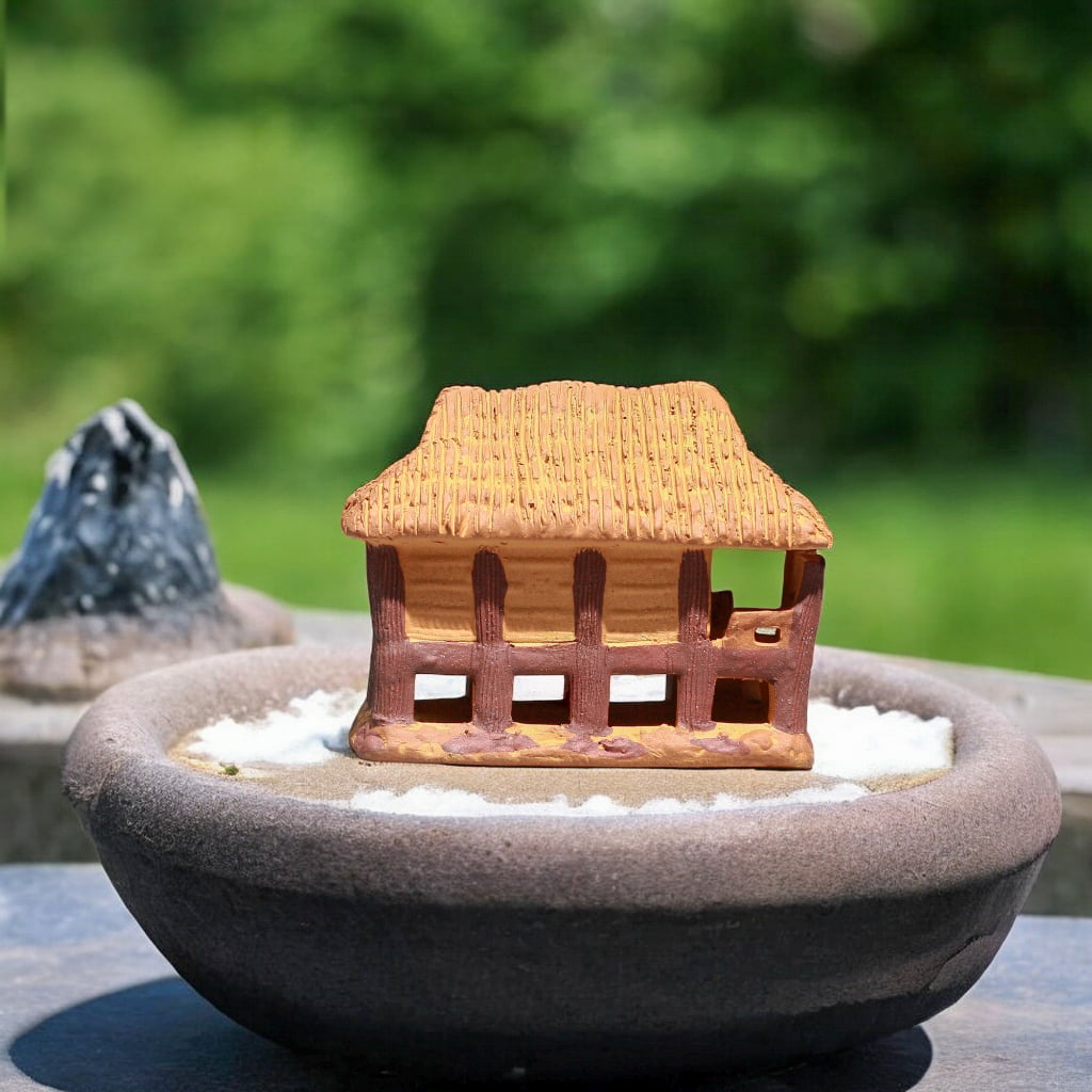 Brown miniature stilt house figurine sits in dark bonsai pot with white sand.