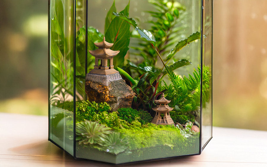 A glass terrarium with two small ceramic pagoda figurines on a mossy hill, surrounded by plants.