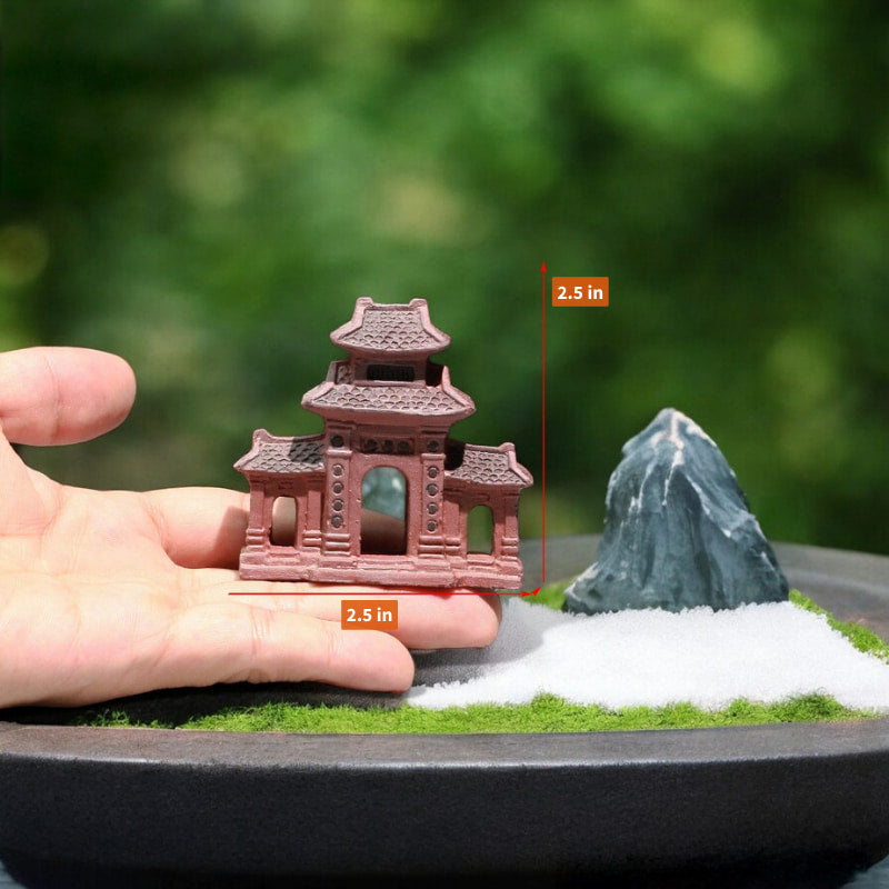 A small brown three entrance gate statue is held in a hand, next to a rock and white sand.