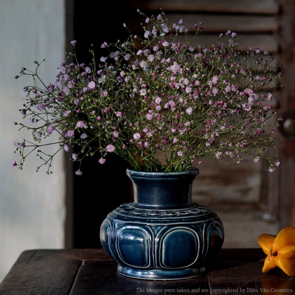 A dark blue artisan ceramic vase holds baby's breath. A Vietnamese ceramic vase on a dark table.
