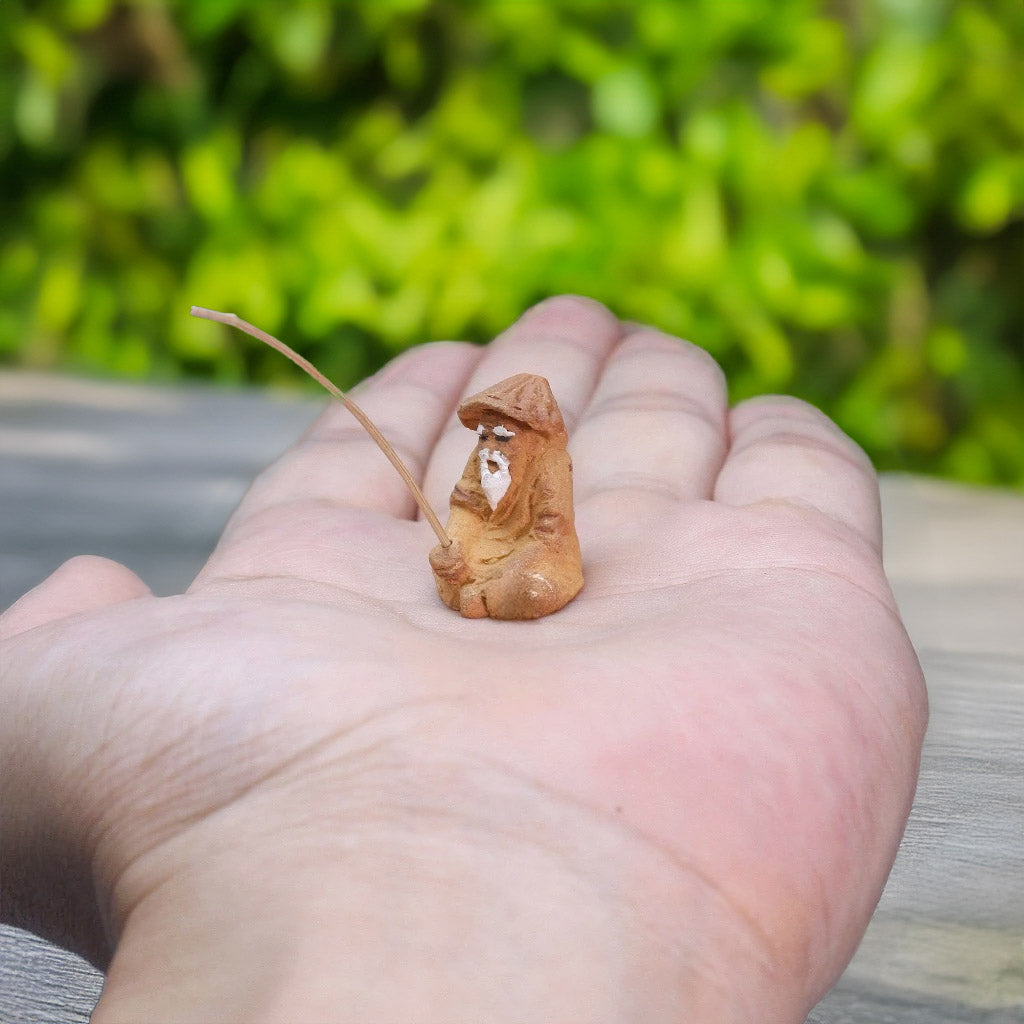 Resting in the palm of a hand, a miniature mudman, with his fishing rod held gently, showcases the rustic charm and intricate detail of this handcrafted figurine.