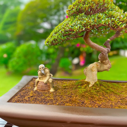 Beneath the shade of a bonsai tree, a miniature Mudman, crafted with intricate detail, sits by his basket trap, adding a touch of Vietnamese tradition to this serene setting.