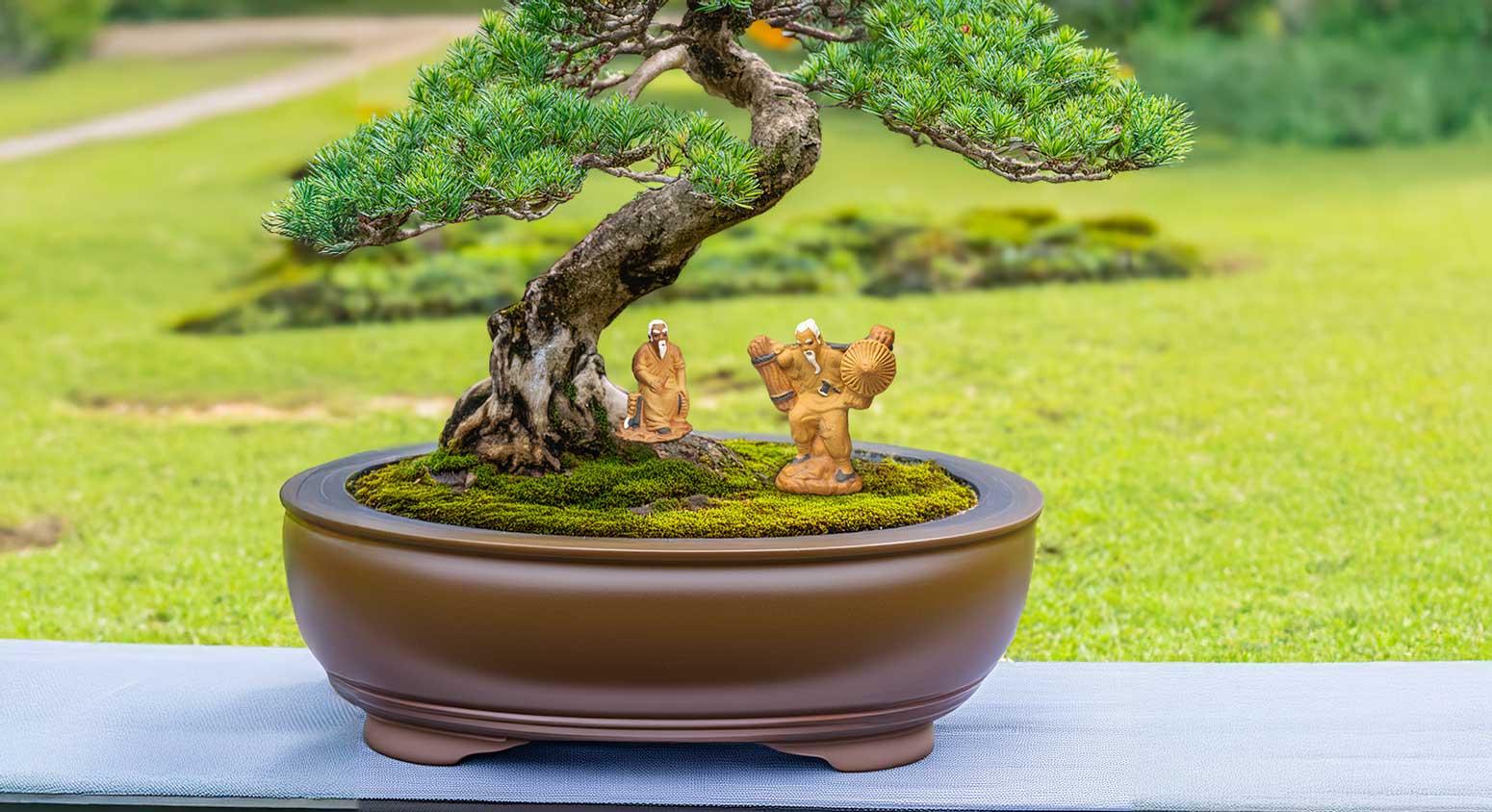 Two miniature ceramic figures, one sitting and one standing, are placed beneath a bonsai tree. The Bonsai figures for bonsai deocration depict woodcutters, one resting and one carrying firewood.