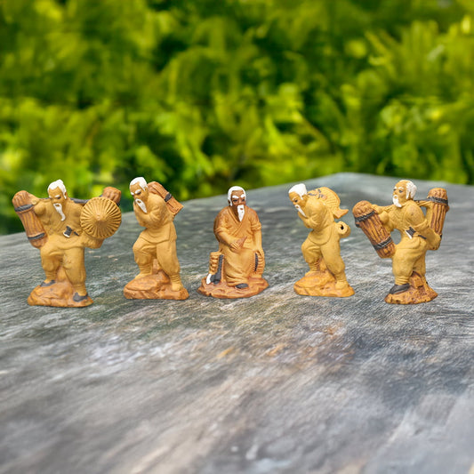 Five small ceramic figures of woodcutters, each with different poses and accessories, are arranged on a wooden surface