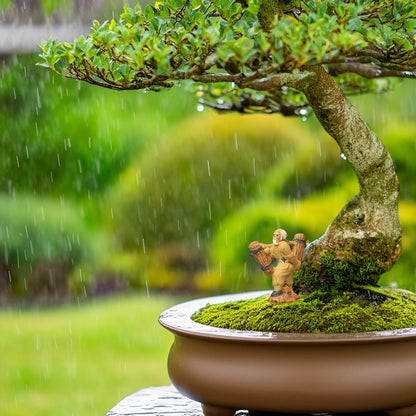 A small ceramic woodcutter stands on a moss-covered mound beneath a bonsai tree. It's raining in the background.
