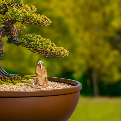 A small ceramic bonsai figure of an old man is sitting on a rock beneath a bonsai tree.
