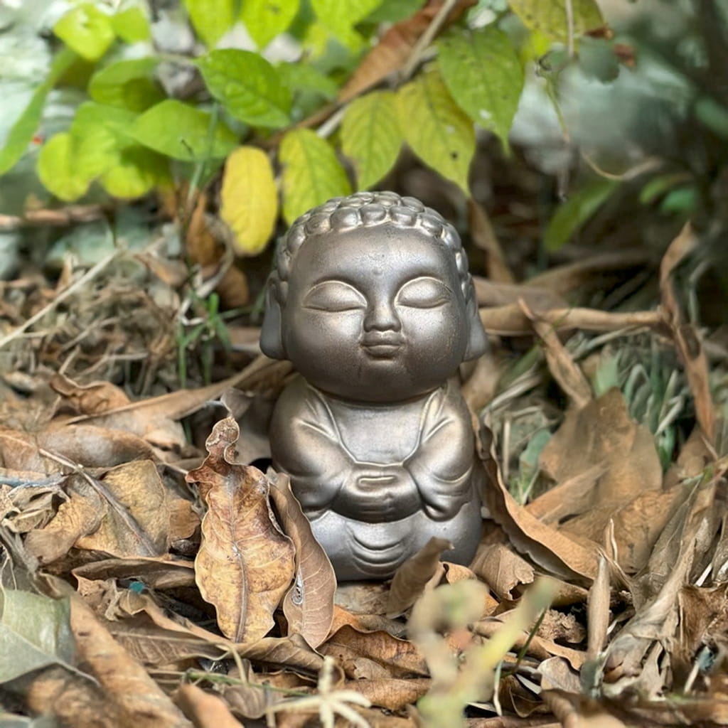 A metallic buddha garden statue rests on dried leaves. A serene bonsai buddha statue for decor.
