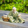 A ceramic mudman with books figurine sits on a wooden surface, ideal for bonsai decor, with a green background.