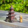 A miniature red pagoda and a mudman figurine are placed on a wooden surface with a green, leafy background. Suitable for Japanese terrarium decor.
