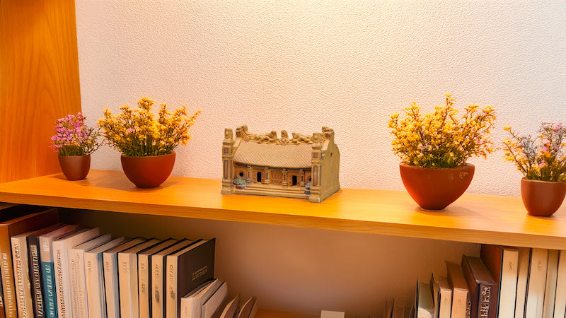  A miniature Vietnamese communal house figurine, crafted from Bat Trang ceramic, sits gracefully on a bookshelf amongst colorful flowers, adding a touch of cultural charm to any space.