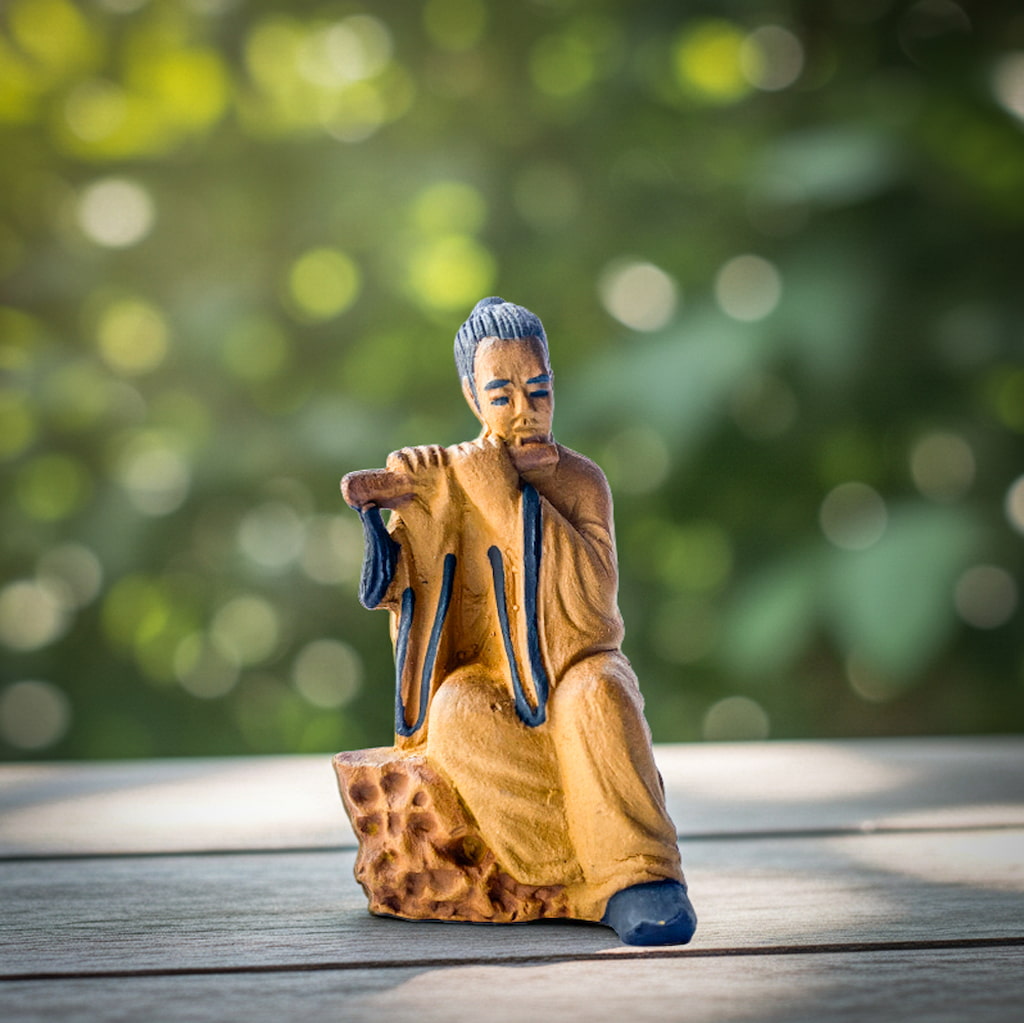 Flute Player Mudmen Figurine Ceramic Sitting Pose on a wooden surface with a blurred green background