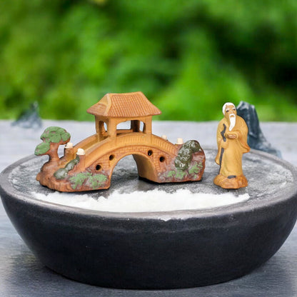 A miniature bridge with a pagoda roof stands on a bed of white sand in a black bowl, a small figurine of an old man with a staff stands beside it, evoking a peaceful Zen garden setting.