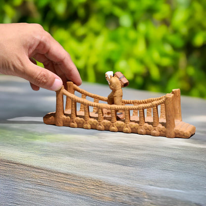 A hand is placing a small ceramic bridge with a rope railing on a wooden surface. A tiny figure of an old man is walking on the bridge.