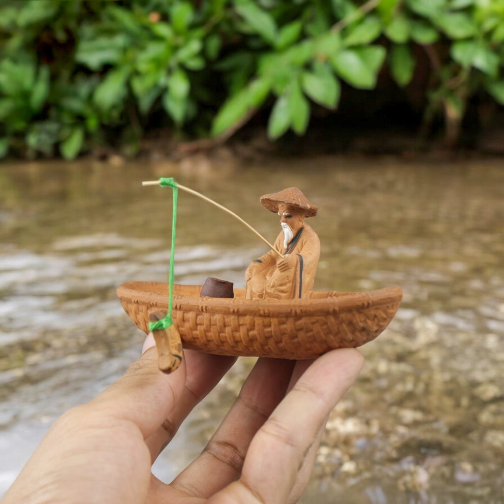 Hand holds small brown fisherman ceramic figurine in boat, rod out. Fishing figurine style.