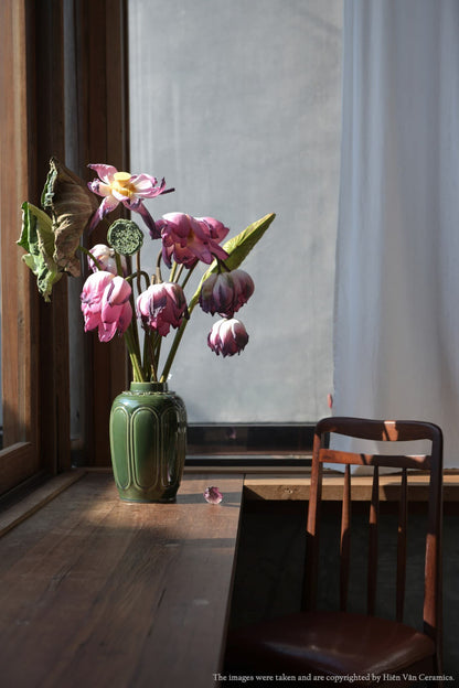 A green Vietnamese ceramic vase with wilting pink lotus on a sunny wooden windowsill.