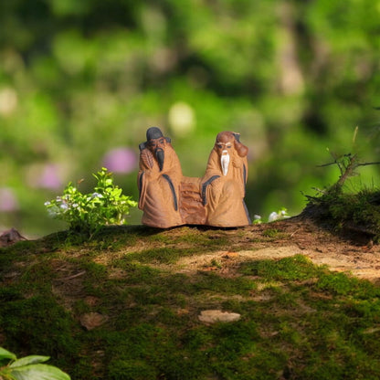 Two small brown ceramic mudmen figurines are sitting on a mossy surface.