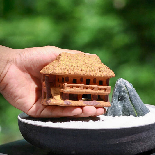 Hand holds vietnamese stilt house figurine over bonsai pot with miniature rock. 