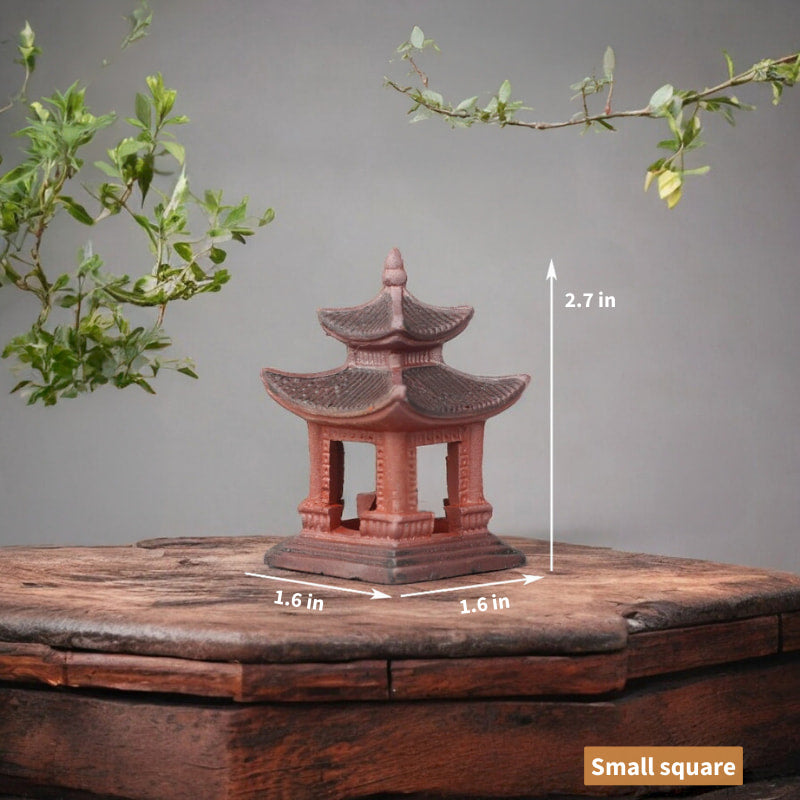 A miniature Japanese pagoda made of red ceramic rests on a rustic wooden table. The pagoda features a square base and two tiers with delicate detailing.