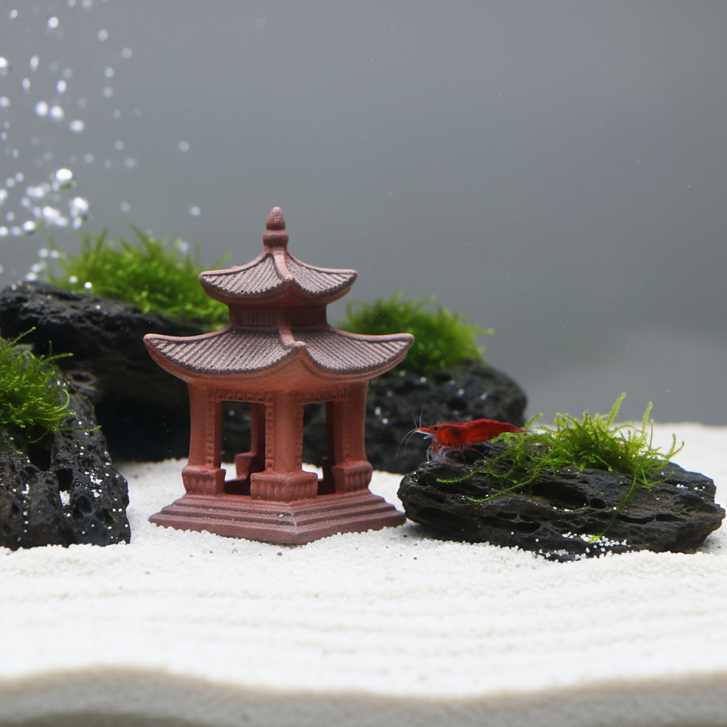A reddish-brown ceramic miniature pagoda sits on white sand in an aquarium. A small red cherry shrimp rests on a nearby moss-covered lava rock. The handcrafted clay structure features detailed tiered roofs and is safe for aquatic life.