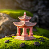 A two-tiered japanese pagoda decor piece with open pillars and a textured grey roof. This ceramic figurine is placed on vibrant green moss, highlighting its rustic reddish clay finish suitable for bonsai figurines.