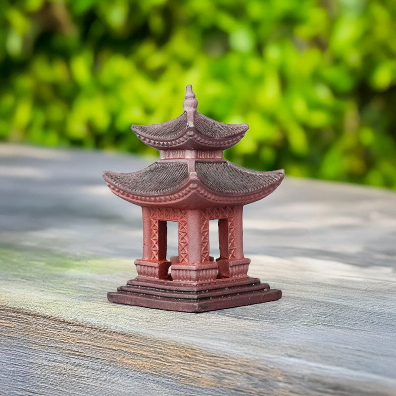 A small, red Japanese pagoda sits on a white sandy bottom in an aquarium. Vibrant green aquatic plants and a piece of driftwood create a natural habitat around the pagoda.