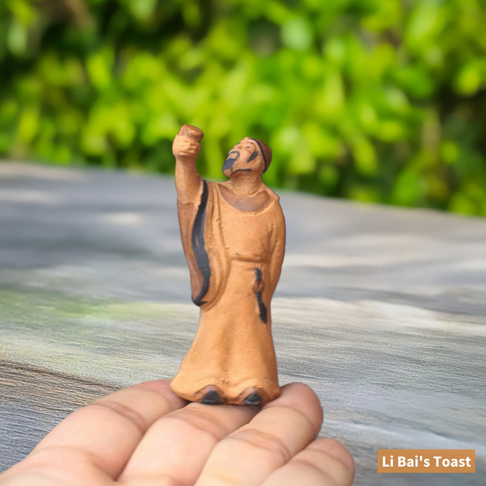 A small, unglazed ceramic figure of Li Bai, a renowned Chinese poet, raising a cup in his hand. He is wearing a long robe and standing on a person's finger.