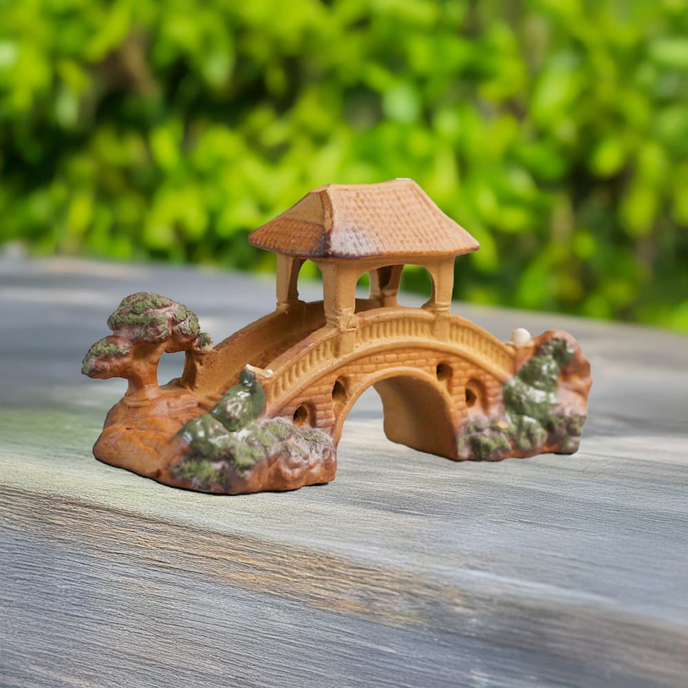 A handcrafted miniature bridge made of brown ceramic, featuring a pagoda-style roof, is displayed on a light wooden surface against a blurred green background.